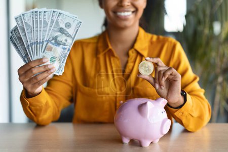 Téléchargez les photos : Successful black businesswoman putting bitcoin coin in piggybank and holding money cash, sitting in office interior. Todays savings are tomorrows money in the bank. - en image libre de droit