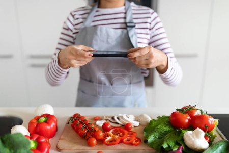 Foto de Female hands holding modern cell phone, cropped of woman in apron taking photo of various fresh organic vegetables on table while preparing healthy meal at home, kitchen interior. Food blogging - Imagen libre de derechos