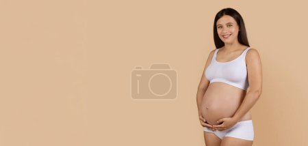 Foto de Pregnant Young Happy Woman In Underwear Touching Her Belly And Smiling At Camera, Beautiful Lady Expecting Baby Caressing Tummy While Posing Over Beige Studio Background, Panorama With Copy Space - Imagen libre de derechos