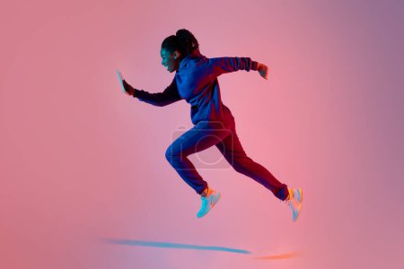 Photo for Full body length shot of modern sporty lady jumping or running using cellphone over pink neon light color background, side view, free space - Royalty Free Image