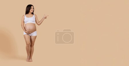 Foto de Great Offer. Happy Young Pregnant Female In Underwear Pointing Aside At Copy Space Over Beige Studio Background, Cheerful Smiling Expectant Woman Demonstrating Free Place For Pregnancy Advertisement - Imagen libre de derechos