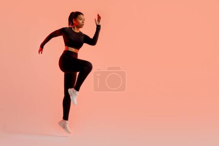 Foto de Sporty black lady jumping, having cardio training, wearing black fitwear, exercising over neon peach studio background, free space. Fitness workout concept. Full length - Imagen libre de derechos