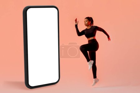 Photo for Workout application concept. Slim lady exercising near big cellphone, jumping in studio over neon peach background. Great mobile offer, gym app concept. Mockup - Royalty Free Image