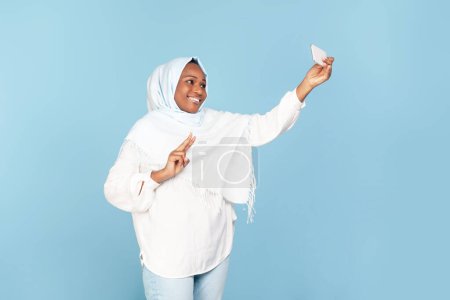Foto de Excited black muslim woman in hijab taking selfie on smartphone and showing peace gesture, making photo standing over blue background, studio shot, copy space - Imagen libre de derechos