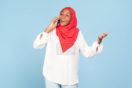 Photo for Emotional african american muslim woman talking on cellphone and gesturing, standing on blue studio background. Happy black lady in hijab enjoying phone conversation - Royalty Free Image