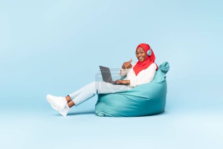 Photo for I like. Happy black muslim woman in headphones sitting on bean bag chair with laptop and showing thumb up, blue background. Lady in hijab working or studying remotely - Royalty Free Image