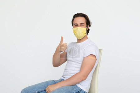 Foto de Smiling young caucasian man in protective mask with band-aid on shoulder after vaccination shows thumb up approves immunization, isolated on white background. Recommendation and advice, health care - Imagen libre de derechos