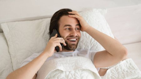 Téléchargez les photos : Cheerful shocked young caucasian guy lies on bed, wakes up, calls by phone, looks at free space in bedroom, top view, panorama. Reaction to good news, win and success, emotions, great ad and offer - en image libre de droit