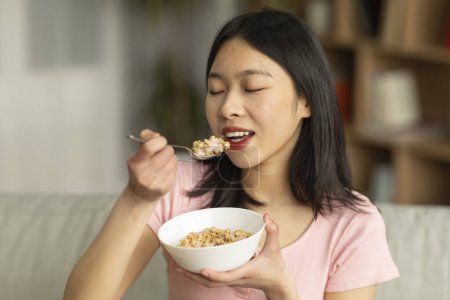 Photo for Portrait of young asian lady having cereal with milk while sitting on sofa at home, free space. Korean woman enjoying healthy meal, keeping balanced weight loss diet - Royalty Free Image