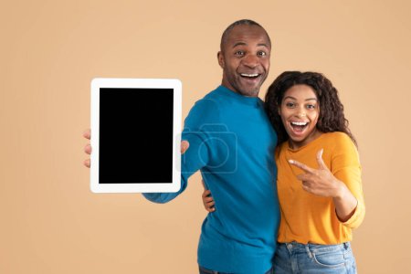 Téléchargez les photos : Happy african american adult couple showing tablet, woman pointing at screen with mockup for design, advertising new app or website, peach background. Touch pad display template - en image libre de droit