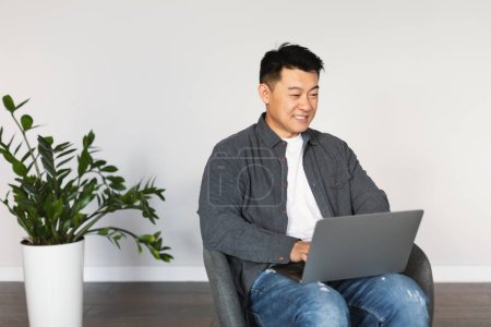 Photo for Smiling middle aged chinese male sitting in armchair, texting, typing on computer, surfing in internet, watch video in living room interior with white wall. Device for business, work remotely at home - Royalty Free Image