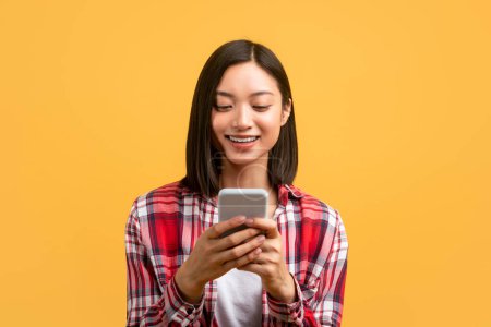 Foto de Young asian woman using smartphone for messaging, smiling lady texting on cellphone while standing over yellow studio background, free space - Imagen libre de derechos