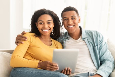 Foto de Cheerful millennial african american man and woman surfing in internet, have meeting on tablet, look at camera in living room interior. Chat and video call, modern device for remote communication - Imagen libre de derechos