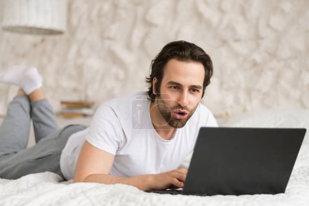 Photo for Glad surprised young caucasian male lies on bed, watching video, searching on computer, has online call in bedroom. Chat in social networks, blogging and surfing in internet, good news, ad and offer - Royalty Free Image