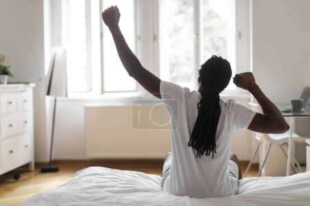 Foto de Rear View Of African American Man Stretching In Bed In The Morning, Unrecognizable Black Male Resting In Light Bedroom After Waking Up, Looking At Window, Feeling Happy After Good Sleep, Copy Space - Imagen libre de derechos