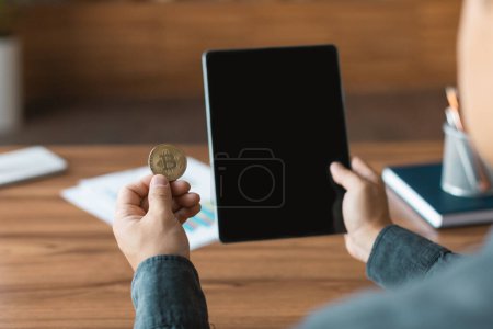 Foto de Adult asian guy looks at tablet with empty screen and bitcoin coin, analyzes financial data at workplace in office interior. Cryptocurrency, economics, saving, app and technology for modern business - Imagen libre de derechos
