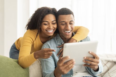 Téléchargez les photos : Cheerful millennial african american lady hug guy, looking at tablet, have video call, use app in living room interior, close up. New normal and lifestyle, technology for communication, ad and offer - en image libre de droit