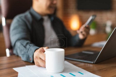Téléchargez les photos : Smiling adult asian guy drinking cup of coffee, typing on smartphone at workplace with laptop in office interior, selective focus, cropped, close up. Break from business, work and startup planning - en image libre de droit