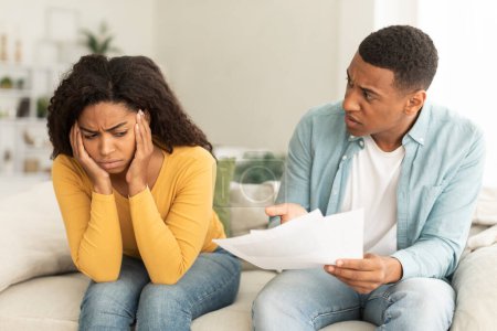 Photo for Angry sad millennial african american man with papers swearing woman due debts, taxes and bills in living room interior. Emotions, problems with finances, bankruptcy, stress and money crisis at home - Royalty Free Image