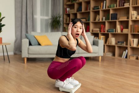 Téléchargez les photos : Shocked asian lady sitting on scales, terrified of weight gain at home, touching head and looking at camera, free space. Woman feeling unhappy with result of her slimming diet - en image libre de droit