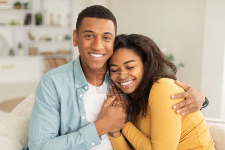 Téléchargez les photos : Cheerful handsome millennial african american male hugging female, enjoy spare time and looking at camera in light living room interior. Love, romantic and family relationship, great mood at home - en image libre de droit