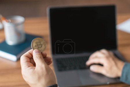 Téléchargez les photos : Middle aged asian man looks at computer with empty screen hold bitcoin, analyzes financial data for investment at workplace in office interior. Cryptocurrency, economics, tech for modern business - en image libre de droit