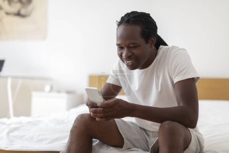 Téléchargez les photos : Smiling Young Black Man Messaging On Smartphone While Relaxing In Bedroom At Home, Happy African American Male Sitting On Bead And Using Mobile Phone For Online Coomunication, Copy Space - en image libre de droit