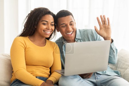 Téléchargez les photos : Happy millennial african american wife and husband looking at laptop, have video call, waving hand in living room interior. Greeting, say hello, hi, meeting remotely and communication remote at home - en image libre de droit