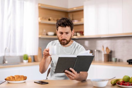 Téléchargez les photos : Serious concentrated millennial caucasian guy with beard in domestic clothes drinks, has breakfast, coffee and reads book at table in kitchen interior. Hobby, literature, study and knowledge at home - en image libre de droit