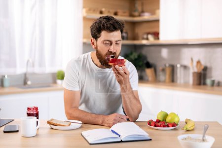 Téléchargez les photos : Serious millennial caucasian guy with beard eats sandwich with jam, reads book at table in kitchen interior. Breakfast at home with literature, planning day with notepad, good morning, time management - en image libre de droit