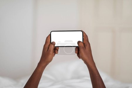 Téléchargez les photos : Mobile Gaming. Unrecognizable Black Male Using Blank Smartphone With White Screen While Lying In Bed At Home, African American Man Playing Online Games While Resting In Bedroom, POV Shot, Mockup - en image libre de droit