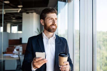 Téléchargez les photos : Confident middle aged businessman in formal wear with coffee to go and smartphone standing near window in office interior. Man chatting with someone, having break after successful business meeting - en image libre de droit