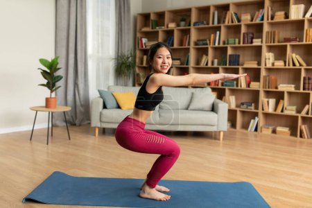 Photo for Happy fit japanese lady in sportswear exercising in living room alone, doing squats, working on buttocks, looking at camera and smiling, side view. Sport at home - Royalty Free Image
