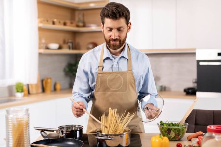 Téléchargez les photos : Smiling attractive millennial caucasian bearded guy in apron cooks spaghetti at table with vegetables in kitchen interior. Prepare dinner for family, hobby and lifestyle at home, household chores - en image libre de droit