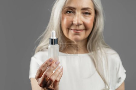 Téléchargez les photos : Cheerful pretty mature european lady shows jar of oil or serum for face, recommend anti-wrinkle cosmetic isolated on gray background, studio. Skin care, anti-aging treatment, spa, rejuvenation at home - en image libre de droit