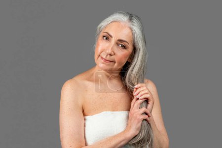 Téléchargez les photos : Serious retired caucasian lady in towel after shower touch gray hair, isolated on gray background, studio, free space. Beauty care, professional treatment, spa beauty day and menopause, ad and offer - en image libre de droit