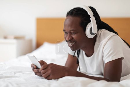 Téléchargez les photos : Smiling Young Black Man In Wireless Headphones Resting In Bed With Smartphone, Happy African American Guy Messaging On Mobile Phone And Listening Favorite Music While Relaxing In Bedroom At Home - en image libre de droit