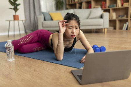 Téléchargez les photos : Tired fit chinese lady lying on yoga mat in front of laptop and wiping her forehead, taking break after intensive home workout. Online domestic sports concept - en image libre de droit