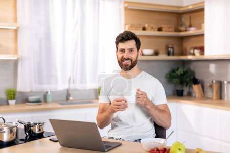 Téléchargez les photos : Smiling millennial caucasian male with beard with cup of coffee enjoys good morning, has breakfast alone with computer in kitchen interior. Weekends and good mood in free time, news and work at home - en image libre de droit