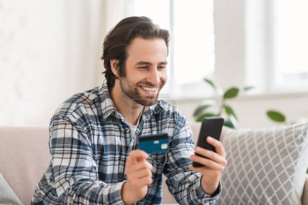 Téléchargez les photos : Cheerful young caucasian guy shopaholic sits on sofa with smartphone and credit card, shopping online, checks financial account in room interior. App for banking, sale and ordering goods remotely - en image libre de droit