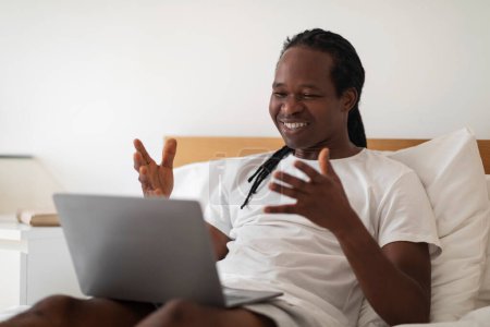 Photo for Video Call. Happy Black Man Making Teleconference Via Laptop While Relaxing In Bed At Home, Cheerful Young African American Guy Talking And Gesturing At Webcamera, Enjoying Online Communication - Royalty Free Image