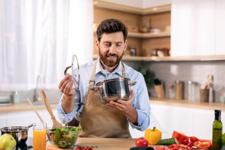 Photo for Happy millennial caucasian bearded male chef in apron prepare food, looks at pan at table with vegetables and salad in kitchen interior. Cooking dinner for family with new recipe, food blog at home - Royalty Free Image
