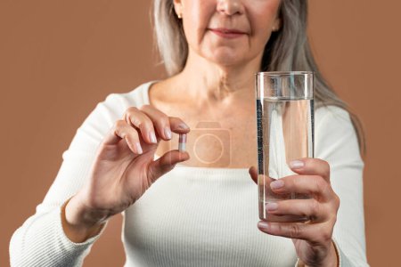Foto de Happy elderly caucasian woman with gray hair shows glass of water and capsule with vitamins isolated on brown background, studio. Beauty care, medicine for hair and skin, antibiotics and pain relief - Imagen libre de derechos