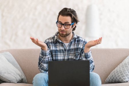 Foto de Surprised confused young caucasian guy blogger in glasses sits on sofa with computer, has video call, gesturing in room interior. Meeting, work remotely, new normal and study, lesson, webinar at home - Imagen libre de derechos