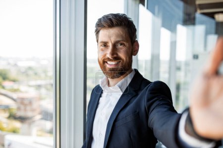 Photo for Happy middle aged businessman having video call or taking selfie while working in office interior and smiling at camera. Confident male entrepreneur communicating online at workplace - Royalty Free Image