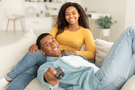 Téléchargez les photos : Cheerful millennial african american male with remote control and female enjoy free time, relaxing on sofa and watching film in light living room interior. Love, romance and movie evening at home - en image libre de droit