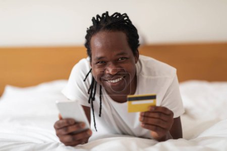 Téléchargez les photos : Online Shopping. Smiling Young Black Man Using Smartphone And Credit Card While Resting In Bed At Home, Happy African American Male Making Purchases From Internet While Relaxing In Bedroom, Closeup - en image libre de droit