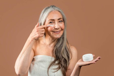 Foto de Smiling mature caucasian woman with natural beauty shows jar and applies cream on face, recommend rejuvenation and moistening at home isolated on brown background. Beauty care, anti-aging treatments - Imagen libre de derechos