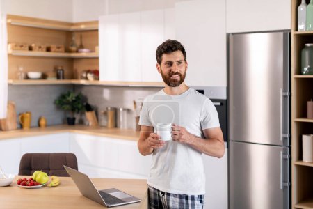 Foto de Happy millennial caucasian guy with beard with cup of coffee enjoys good morning, has breakfast with computer, looks at copy space in kitchen interior. Free time, device for work at home. ad and offer - Imagen libre de derechos