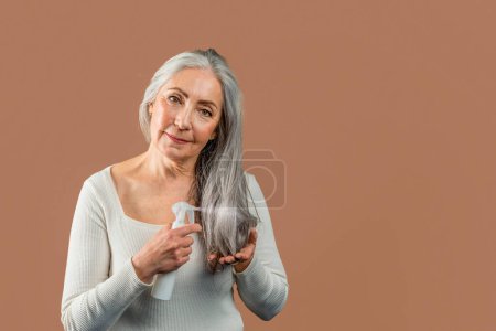 Foto de Smiling mature caucasian female with natural beauty applies spray on gray hair, looking at camera, isolated on brown background, studio. Beauty care, daily treatments for rejuvenation, ad and offer - Imagen libre de derechos